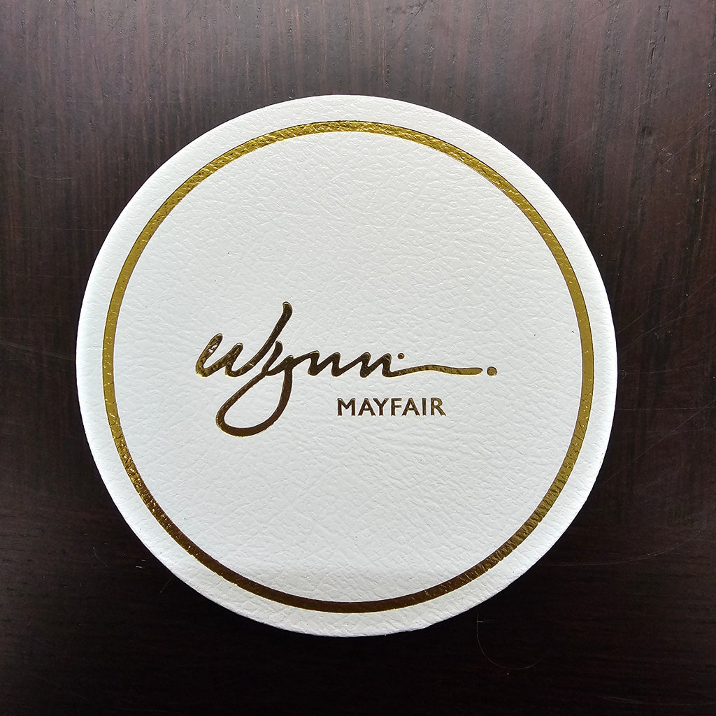 Round white coaster with gold border and 'Mayfair' branding on a dark wooden surface