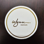Round white coaster with gold border and 'Mayfair' branding on a dark wooden surface